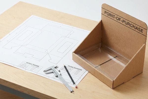 A Point-Of-Purchase cardboard display box prototype with clear tray, dieline blueprint, and caliper for structural packaging.