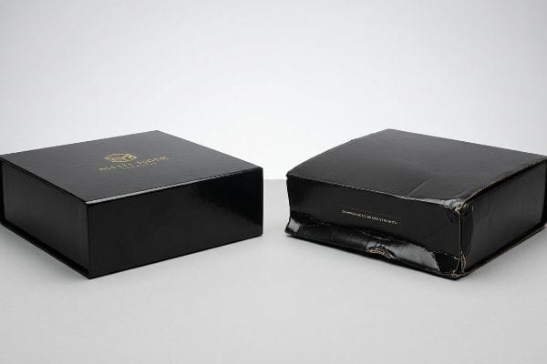 Glossy black rigid box with gold 'Miel & More' logo next to a severely crushed black corrugated box, illustrating transit damage.