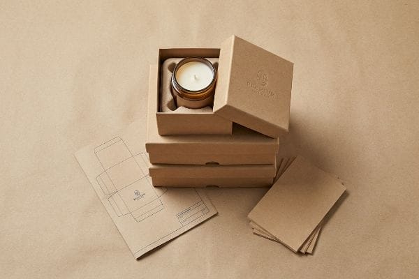An open kraft candle box, embossed 'Premium', displays an amber glass jar candle, with stacked boxes, a flat template, and kraft cards.
