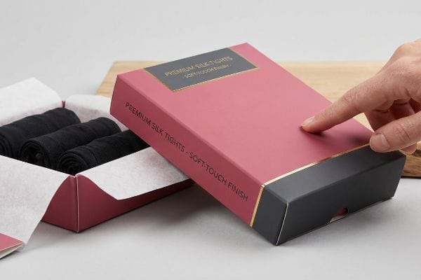Premium Silk Tights Box A hand touches a pink and black box of Premium Silk Tights with Soft-Touch Finish, next to an open box revealing three black tights.