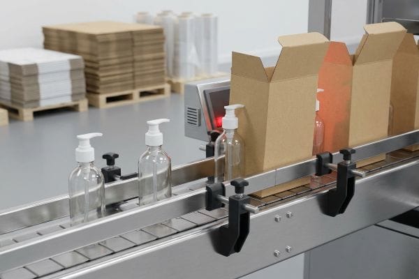 Lotion Bottle Packaging Line Clear plastic pump bottles move along a stainless steel conveyor belt towards open brown corrugated boxes in a packaging facility.