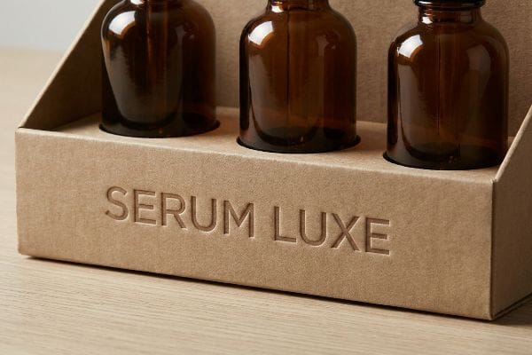 Serum Luxe Debossed Packaging Kraft paperboard tray with debossed 'Serum Luxe' text, holding three amber glass serum bottles for luxury skincare.