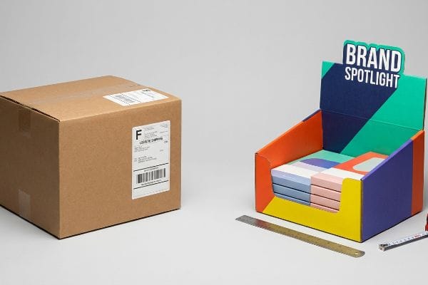 Shipping vs Retail Packaging Brown cardboard shipping box with Logistic Shipping label beside a colorful Brand Spotlight retail display holding pastel product boxes.