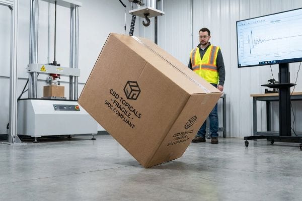 Cbd Drop Test Brown cardboard master carton, labeled 'Cbd Topicals - Fragile - Sioc Compliant', suspended for kinetic drop testing in a packaging fulfillment lab, with a technician observing.