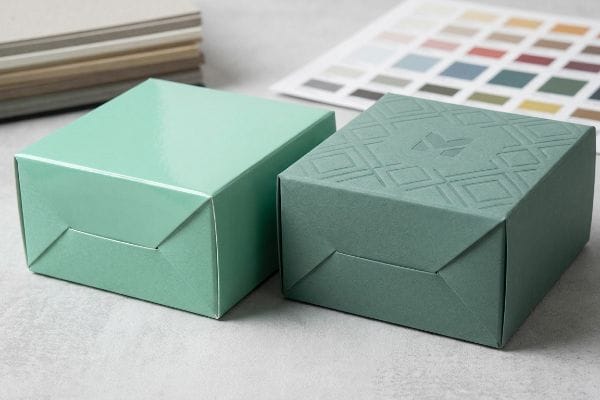 A caixa de presente verde menta brilhante contrasta com a caixa azul-petróleo com textura suave em relevo, destacando os acabamentos táteis.