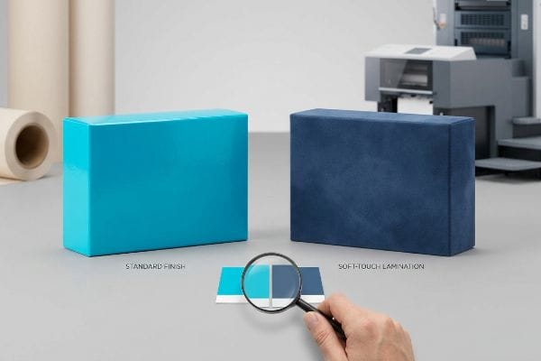 Lamination Color Shift Glossy turquoise Standard Finish box contrasts with velvet navy Soft-Touch Lamination, demonstrating color shift on a swatch with a magnifying glass.
