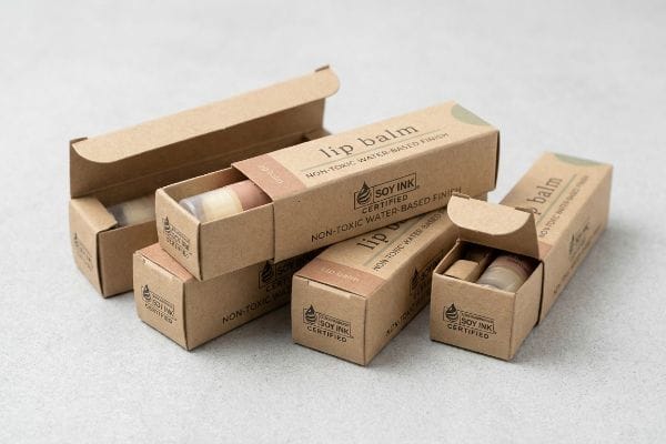 リップクリーム用のクラフト紙製折りたたみ式カートン。化粧品パッケージとして、「大豆インク認証済み」および「無毒性水性仕上げ」の表示が目立つように記載されている。.