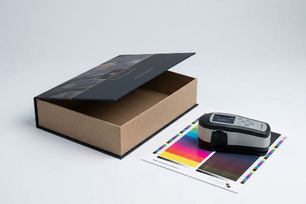 Color Measurement Gift Box An open kraft gift box with a dark laminated lid, next to a spectrophotometer measuring a color calibration chart showing Delta E.