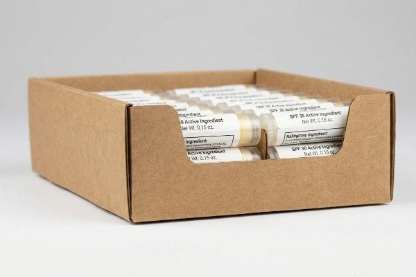 Brown corrugated cardboard display tray holds clear lip balm tubes with visible "SPF 30 Active Ingredient" and "Net Wt. 0.15 oz." labels.