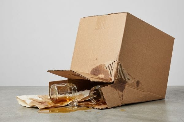 Spilled Liquid Box Collapse Brown cardboard box collapsed from spilled amber liquid, dissolving adhesives, with two glass jars on stained paper.