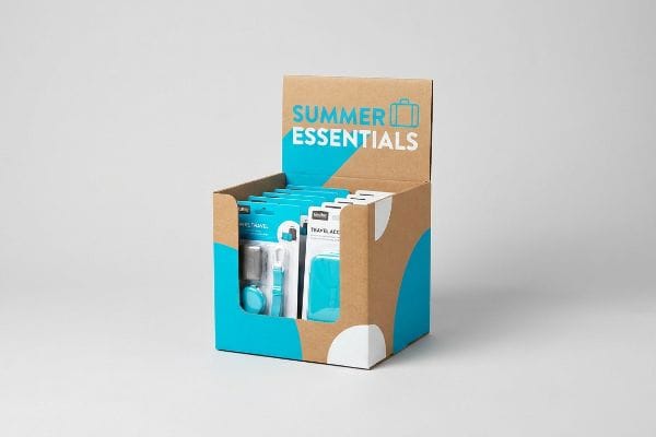 Corrugated cardboard retail display box, teal and white, features "Summer Essentials" with suitcase icon, holding blister-packed travel accessories.