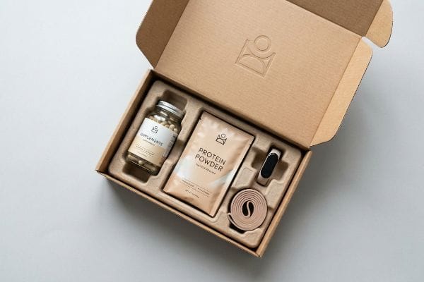 An open health subscription box displays a glass jar of Supplements, Protein Powder pouch, fitness tracker, and resistance band in Kraft paper packaging.