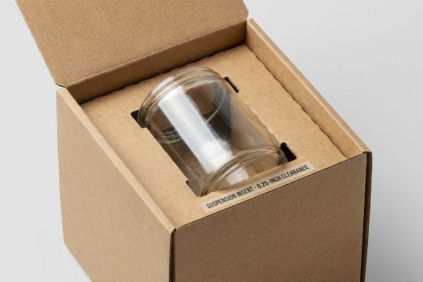 Brauner Wellpappkarton mit einem durchsichtigen Glasgefäß, das von einem ausgestanzten Kartoneinsatz gehalten wird, der mit „Aufhängungseinsatz – 0,25 Zoll Abstand“ beschriftet ist.