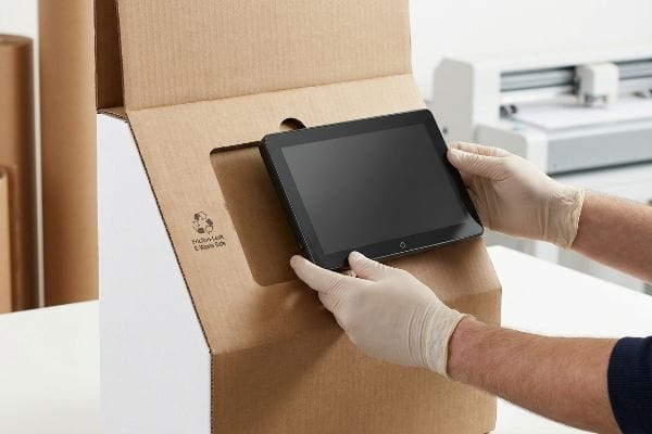 टैबलेट फ्रिक्शन-लॉक बॉक्स "फ्रिक्शन-लॉक ई-वेस्ट सेफ" के रूप में चिह्नित एक नालीदार कार्डबोर्ड बॉक्स में, दस्ताने पहने हाथों से एक काली गोली को उसके कटे हुए खांचे में डाला जाता है।.