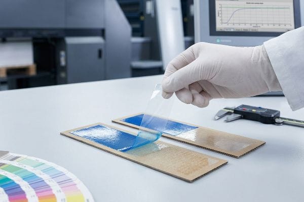 Tape Adhesion Test A gloved hand performs a 3M tape-adhesion test, peeling a clear film from a cardboard strip with a blue coating to check for flaking, ensuring quality control.