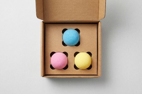 Colorful Bath Bomb Box An open corrugated cardboard bath bomb display box with a custom-engineered die-cut insert cradling three spherical bath bombs: blue, pink, and yellow.