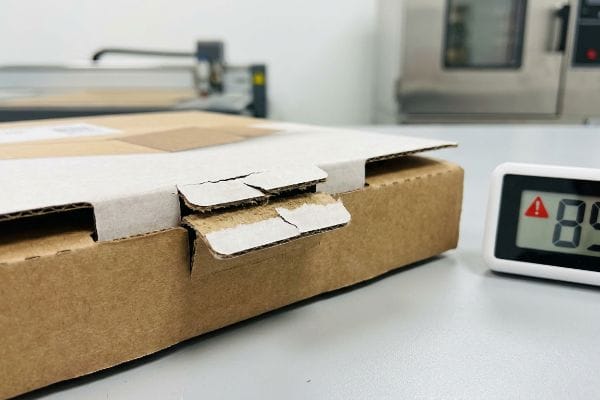 Caja de cartón corrugado marrón con la pestaña de cierre rota, junto a una pantalla digital que muestra un 85% de humedad relativa, lo que ilustra la hinchazón causada por la humedad.