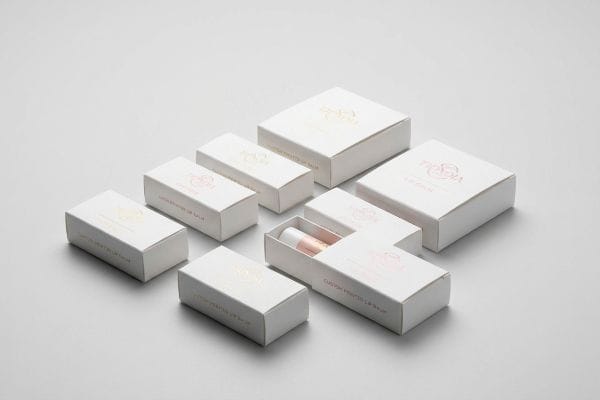 Arranged on a white surface, custom printed Trisha lip balm boxes feature gold and pink foil logos, with one box revealing a lip balm tube.