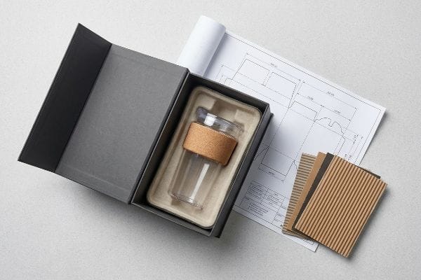 Overhead view of custom tumbler packaging design, featuring a glass tumbler in a molded pulp insert, dieline blueprint, and corrugated samples.