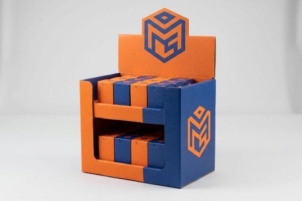 Orange and blue corrugated cardboard two-tiered display stand with a stylized M logo, holding small orange and blue product boxes.
