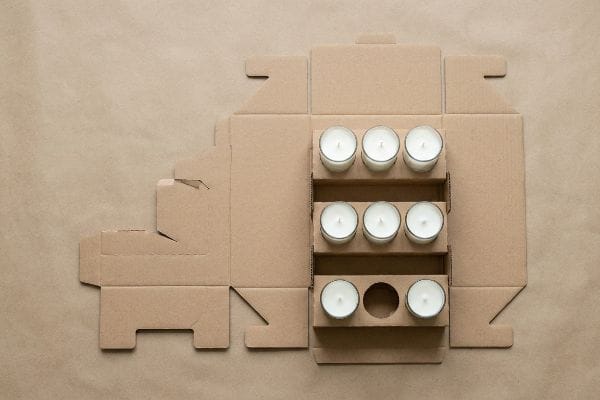 Caja expositora de cartón kraft corrugado desplegada para velas, con nueve velas blancas en portavelas de cristal y separadores internos, lista para su montaje.