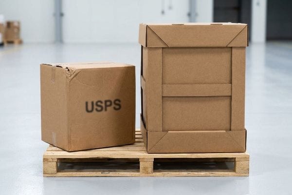 パレットに載せられたUSPSの箱 茶色の段ボール箱(うち1つには「USPS」とラベルが貼られている)が木製パレットの上に置かれており、標準的なサイズの郵便物用ボックスと、より大きな配送用ボックスが並べて展示されている。.