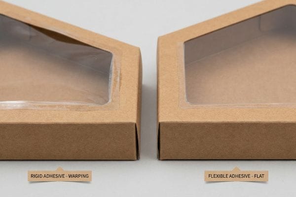 Brown kraft paperboard boxes comparing 'Rigid Adhesive - Warping' with a distorted window patch to 'Flexible Adhesive - Flat' with a smooth window.
