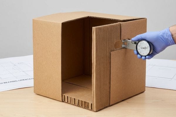 波形フルートの厚さ ノギスで特注箱の段ボールのフルートの厚さを測定し、構造設計の重要性を強調している。.