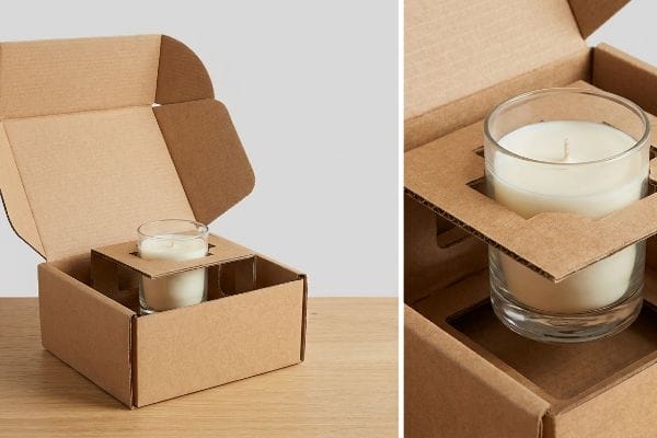 キャンドル用ガラス瓶のパッケージ 茶色の段ボール箱に、特注の型抜きインサートで固定された2つのガラス瓶キャンドルが入っており、輸送用の吊り下げスペースが設けられています。.