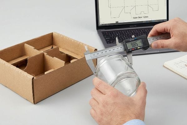 Medición con calibrador de frascos Unas manos miden un frasco de vidrio con un calibrador digital que marca 98,5 mm, junto a una caja de cartón corrugado vacía y un ordenador portátil que muestra el diseño del embalaje Apex Clear de 1,2 mm.
