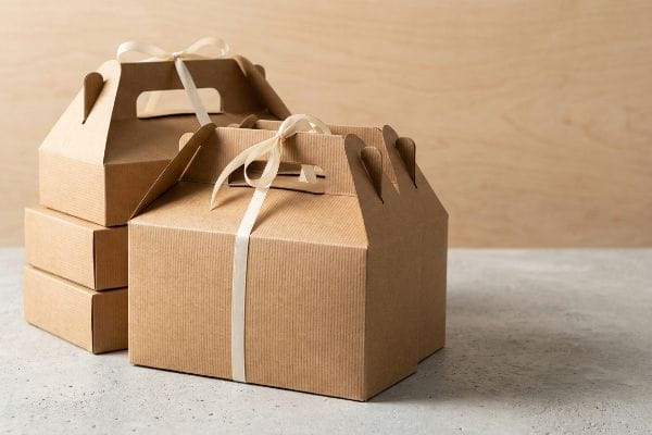 Fitas para caixas Kraft Gable Caixas de cartão kraft, dispostas cuidadosamente e atadas com elegantes fitas creme, assentam sobre uma superfície de betão cinzento sobre um fundo de madeira clara.