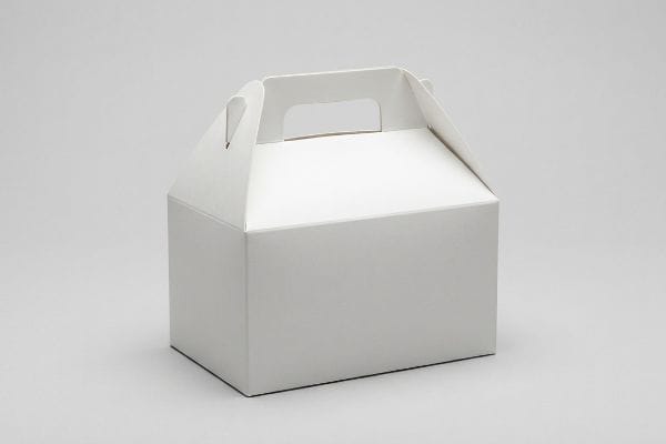 持ち手と傾斜した上部を備えた、ブランド名のない白い厚紙製の三角箱が、薄いグレーの背景に置かれている。.