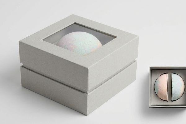バスボムのパッケージディスプレイ 薄いグレーの厚紙製のパッケージには透明な窓があり、パステルカラーのバスボムが1つ、そして半分の大きさのバスボムが2つ入った開いた箱が並んで見える。.