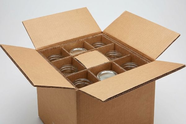 An open corrugated cardboard box with internal dividers securely holding nine clear glass jars, some with silver lids, for fragile item packaging.