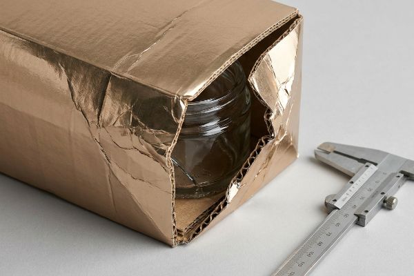 Foil Packaging Caliper Audit Crumpled gold foil lamination on a damaged corrugated box, revealing a glass jar, with a caliper for material tolerance measurement.