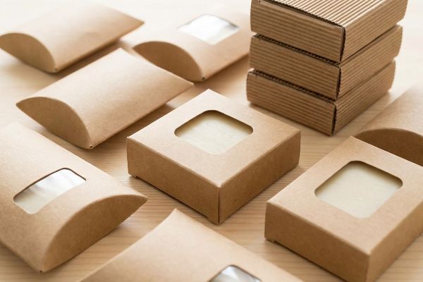 Kraft Window Pillow Boxes Arrangement of unbleached kraft soap boxes, featuring windowed and pillow designs, alongside stacked corrugated packaging.