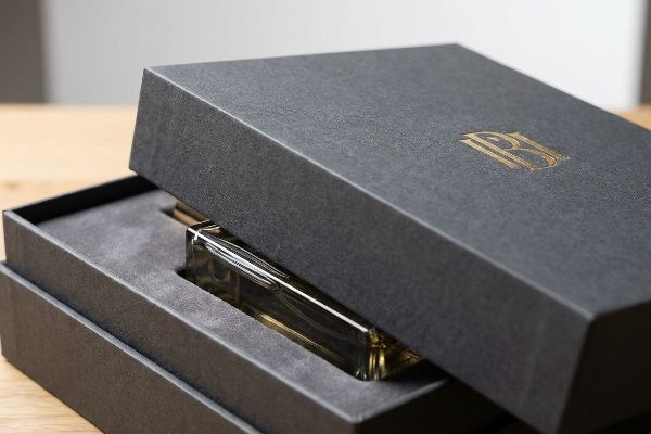 A premium perfume bottle rests in a dark grey textured luxury box with a golden foil RB logo on its partially open lid, showcasing a velvety insert.