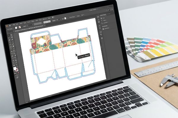 Laptop screen displays a colorful packaging dieline template in Adobe Illustrator, showing a 0.5-inch bleed margin for corrugated box prepress.