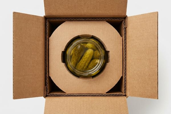 Brown corrugated cardboard box with a glass jar of pickles secured by a die-cut suspension void insert, preventing kinetic shock.