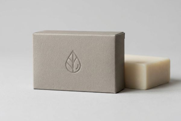 Soft Touch Soap Box Logo Light gray soft-touch paperboard soap box with a debossed leaf droplet logo, beside an off-white natural soap bar.